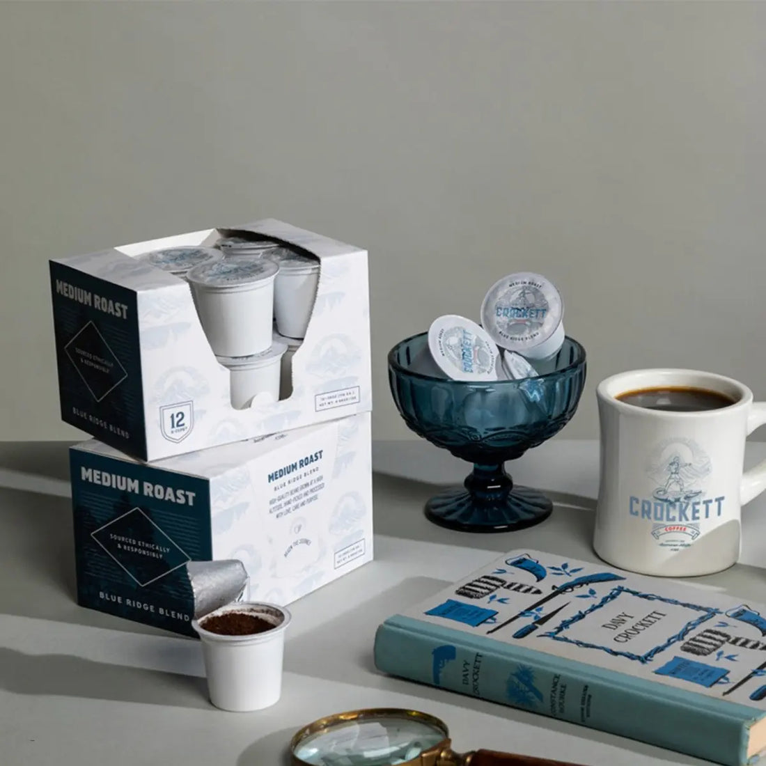 Medium roast coffee pods from Crockett Coffee arranged with a mug, blue glass bowl, and book on a gray surface.
