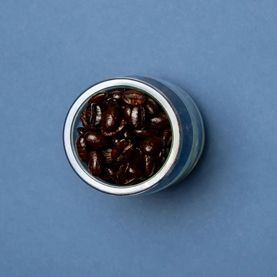 Dark roast coffee beans in a glass jar against a blue background, showcasing their rich, glossy texture