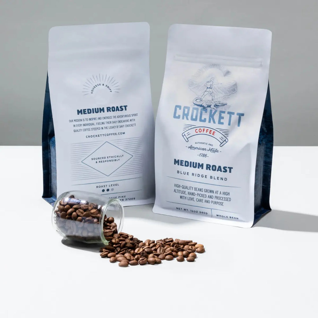 Whole bean coffee in premium packaging with roasted coffee beans spilling from a glass jar onto a white surface.