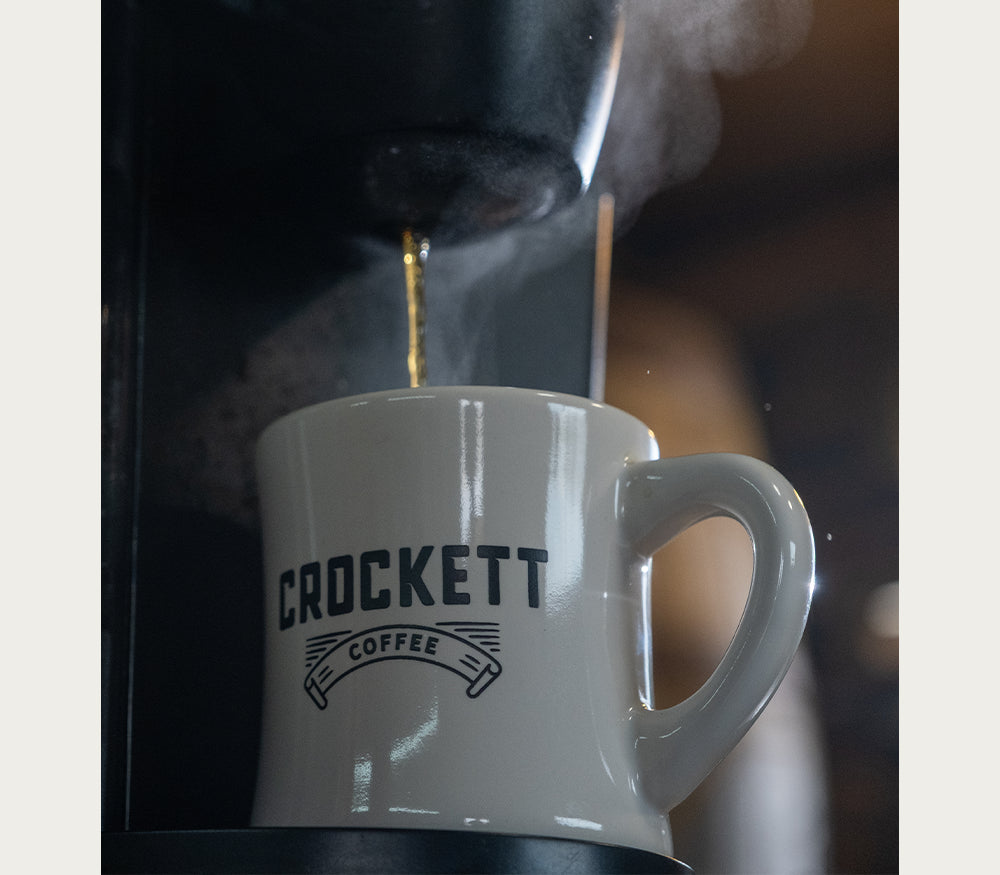 Coffee mug labeled “Crockett Coffee” being filled with fresh hot coffee from a brewing machine.