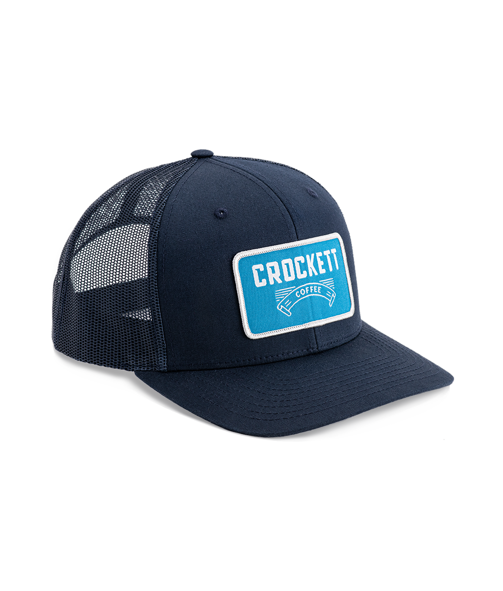 Navy trucker hat with blue Crockett Coffee patch, mesh back, and structured front panel design.