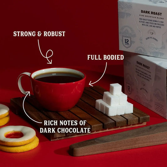 Dark roast organic coffee in a red cup on a wooden coaster with sugar cubes, biscuits, and product boxes on a red background