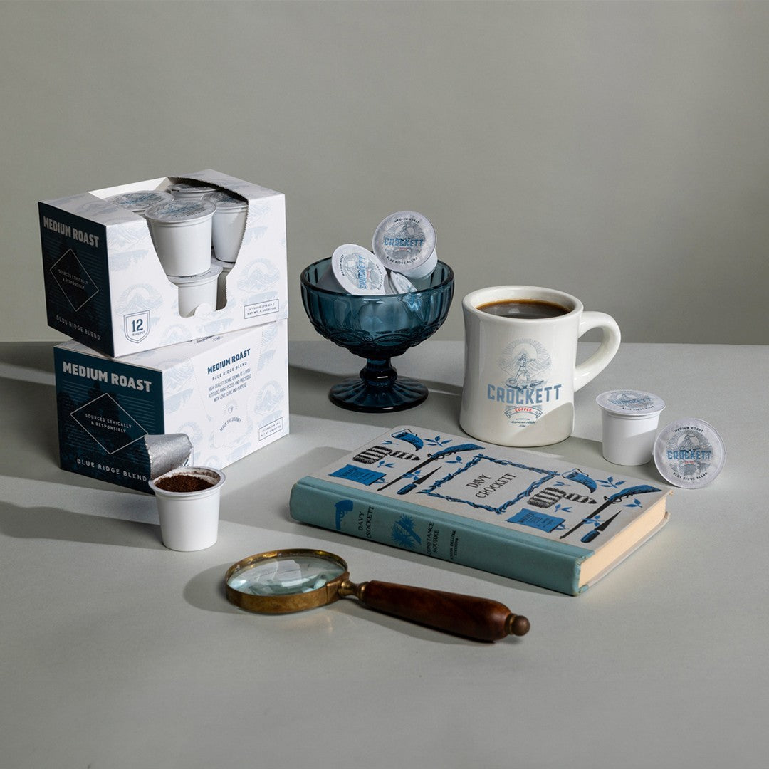 Crockett Medium Roast coffee pods with a branded mug, decorative bowl, magnifying glass, and book on a gray surface