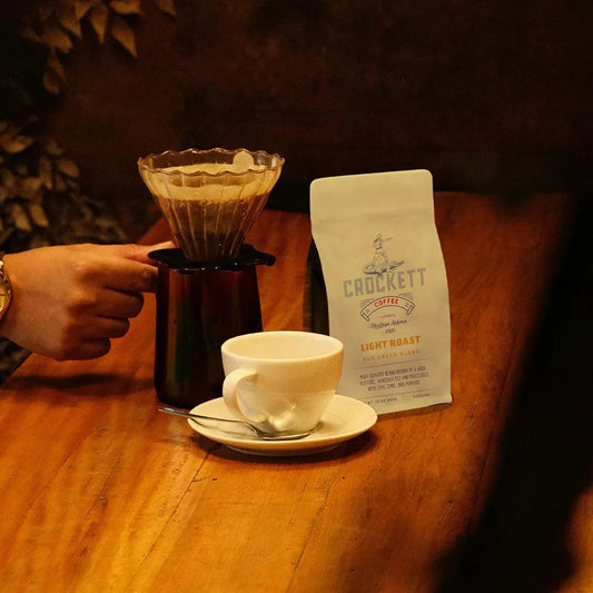 Coffee brewing setup with Crockett Light Roast organic coffee bag, a white cup, and a pour-over filter on a wooden table