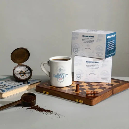 K-cup coffee displayed with a branded mug, chessboard, compass, and scattered coffee grounds on a table.