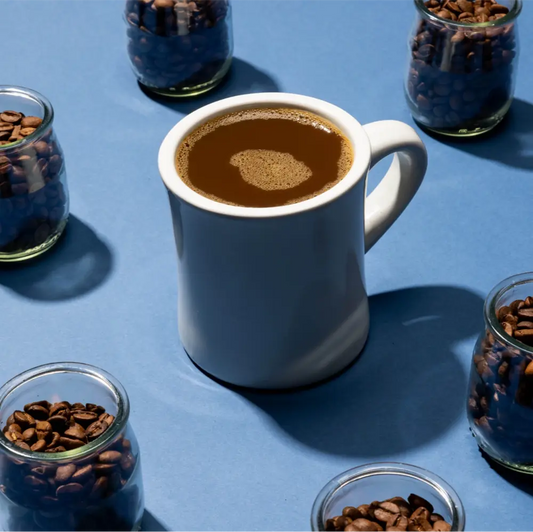 Nitro coffee in a white mug surrounded by glass jars of coffee beans on a bold blue background.