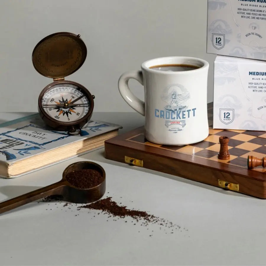 Small-batch coffee mug on a chessboard surrounded by ground coffee, vintage compass, and branded packaging.