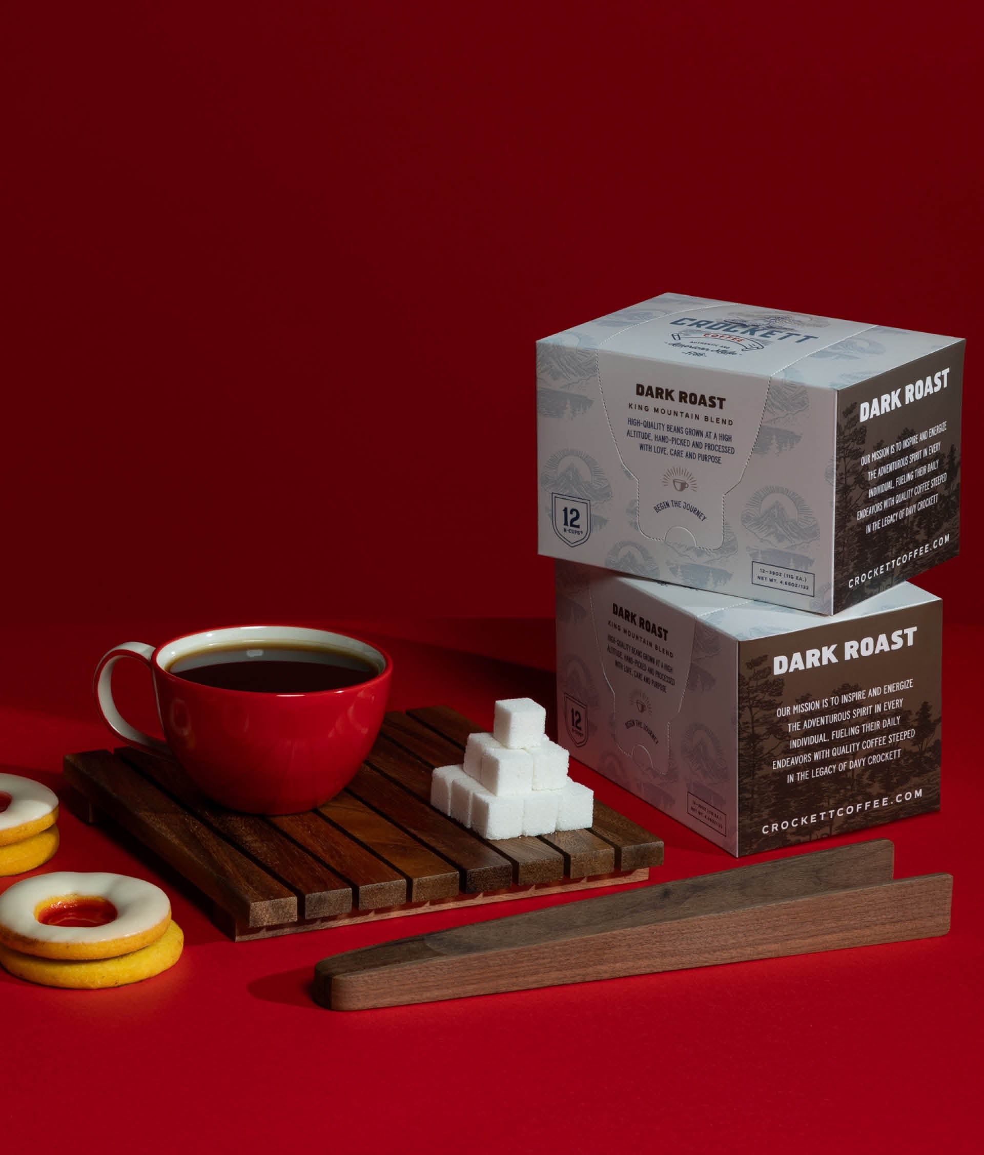 Dark roast coffee K Cups from Crockett Coffee displayed with sugar cubes, cookies, and a red mug on a bold red background.