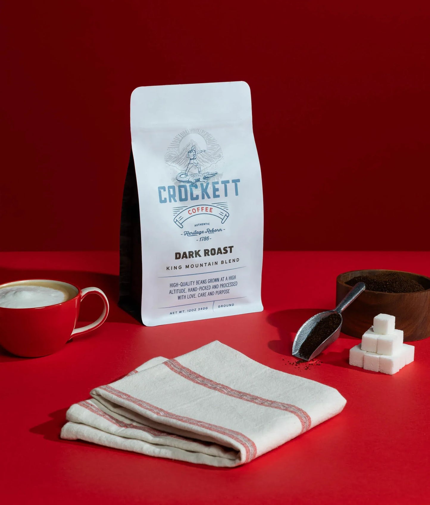 Dark roast coffee bag labeled “King Mountain Blend” from Crockett Coffee, displayed with a red cup of coffee, ground coffee in a scoop, sugar cubes, and a folded napkin on a red background.