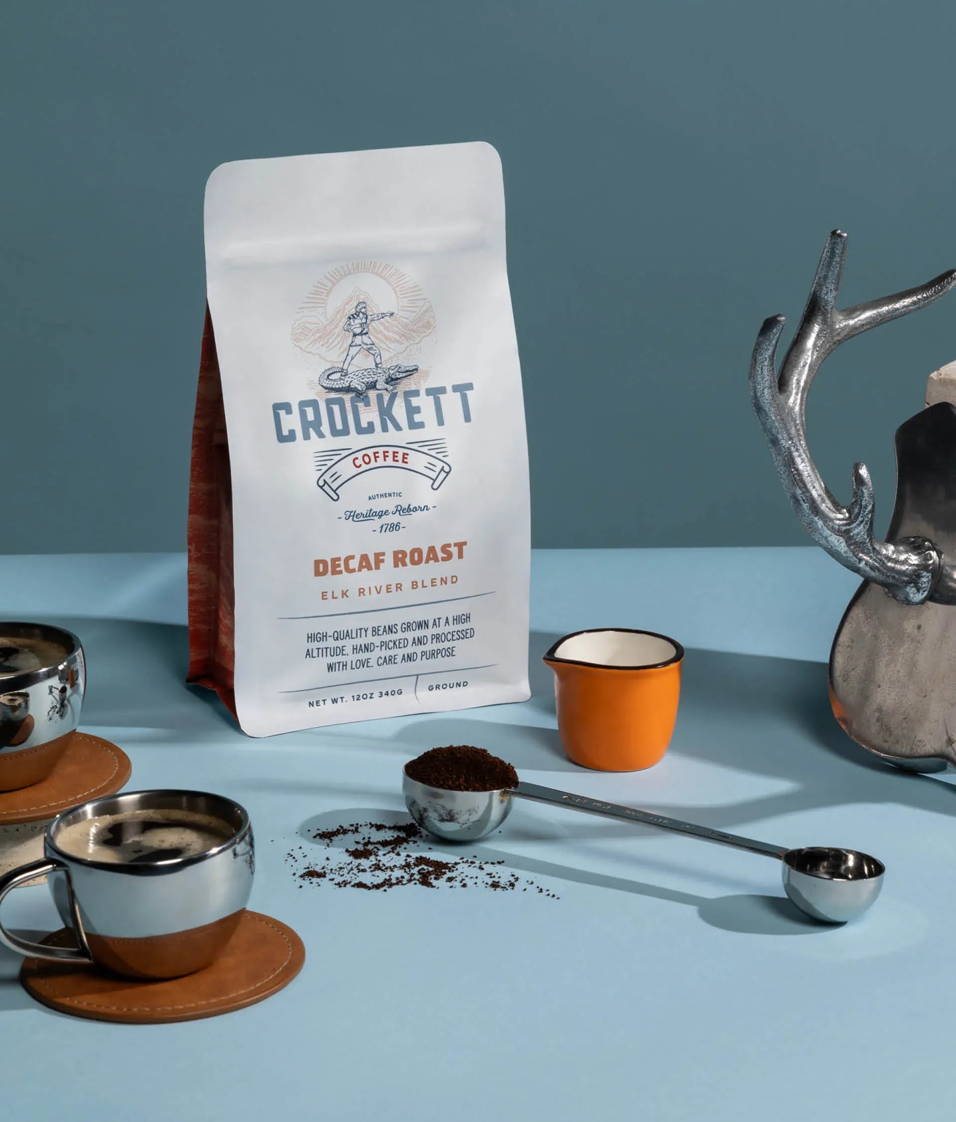 Ground decaf coffee in Crockett Coffee's Elk River Blend bag with a scoop, espresso cups, and rustic decor.