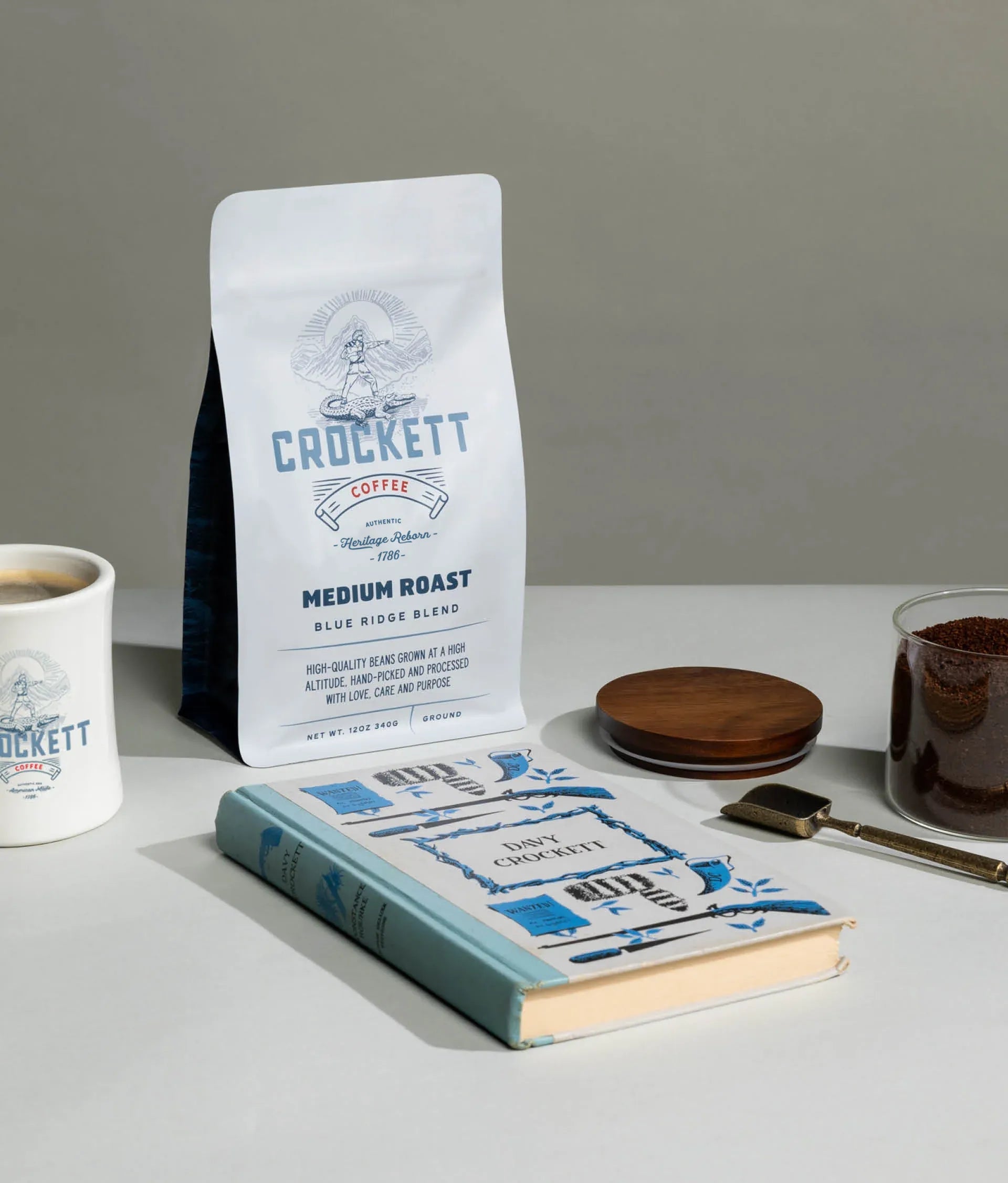 Ground whole bean coffee in Crockett Coffee's Medium Roast Blue Ridge Blend bag beside a mug, book, and coffee grounds.