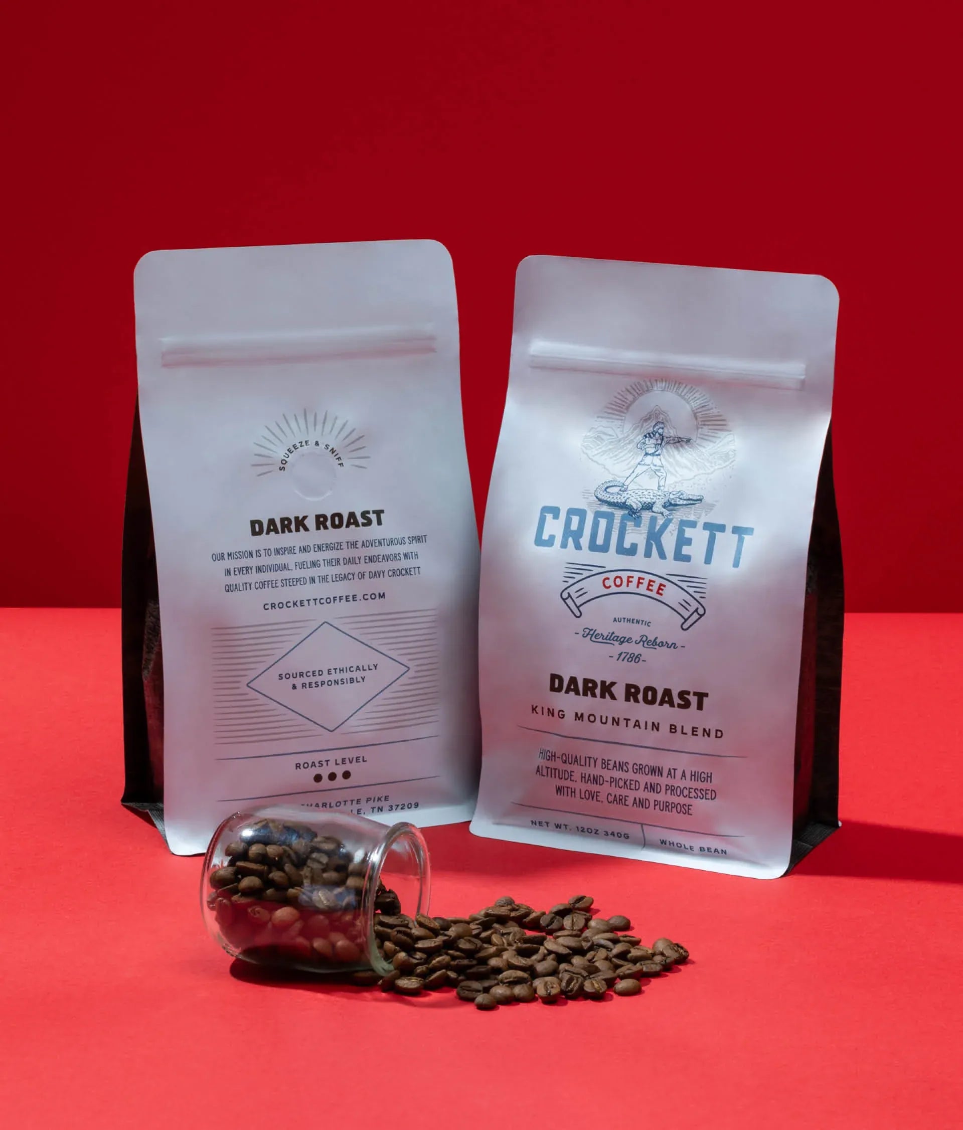 Whole bean dark roast coffee in Crockett Coffee's King Mountain Blend bags with scattered coffee beans on a red background.