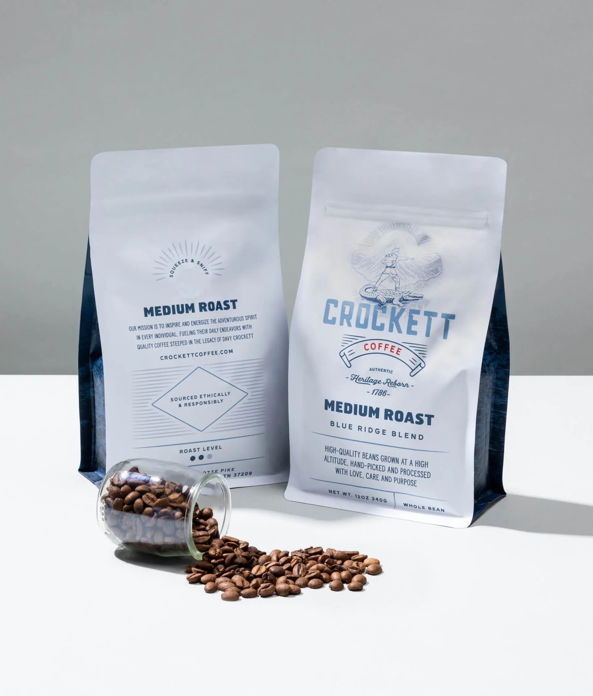 Whole bean medium roast coffee in Crockett Coffee's Blue Ridge Blend bags with spilled coffee beans on a white surface.