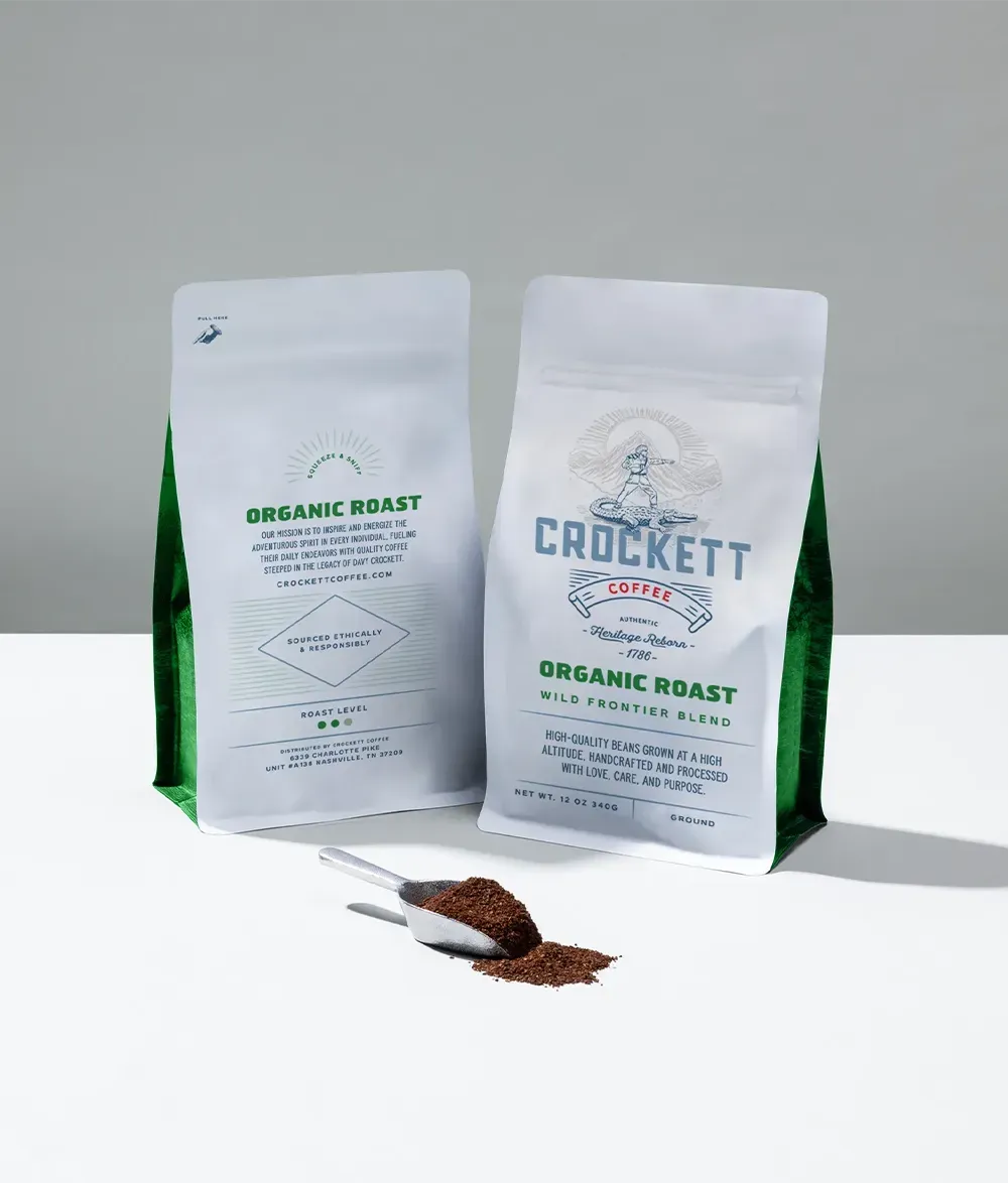 Organic ground coffee in Crockett Coffee's Wild Frontier Blend bags with a metal scoop and loose coffee grounds.