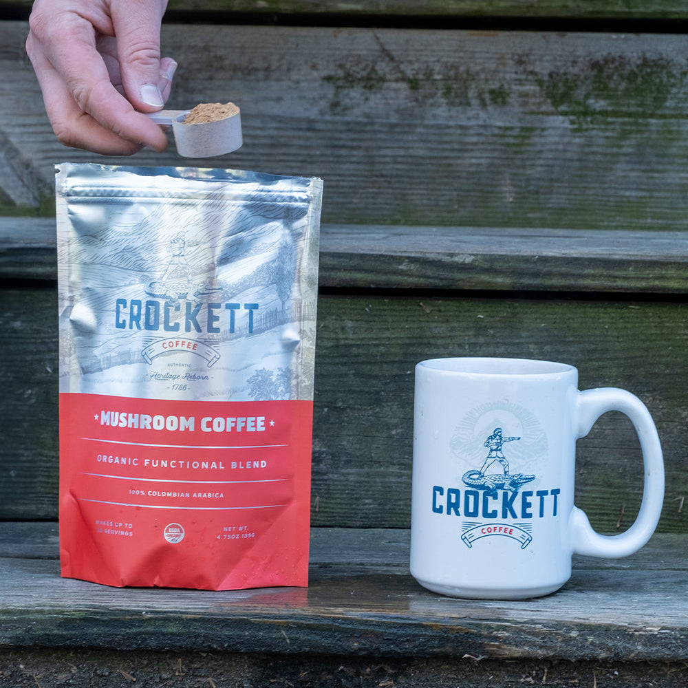 Mushroom coffee being scooped from a Crockett Coffee pouch next to a white branded mug on wooden steps.