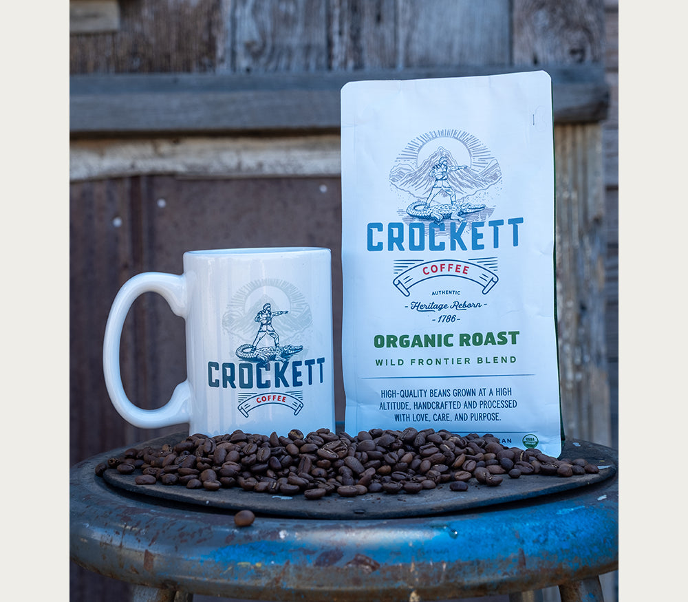 Coffee beans scattered in front of Crockett Coffee organic roast bag and branded mug on a rustic metal table.