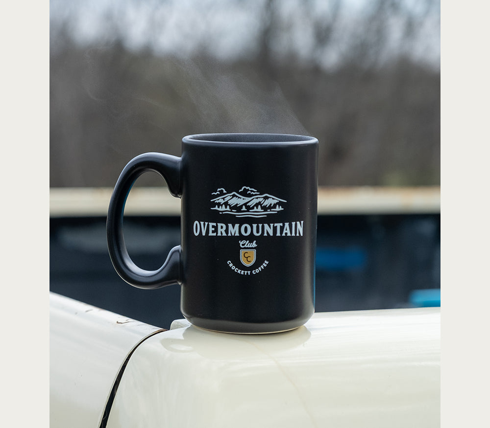 Black coffee mug with Overmountain Club and Crockett Coffee logo steaming atop a white vehicle outdoors.