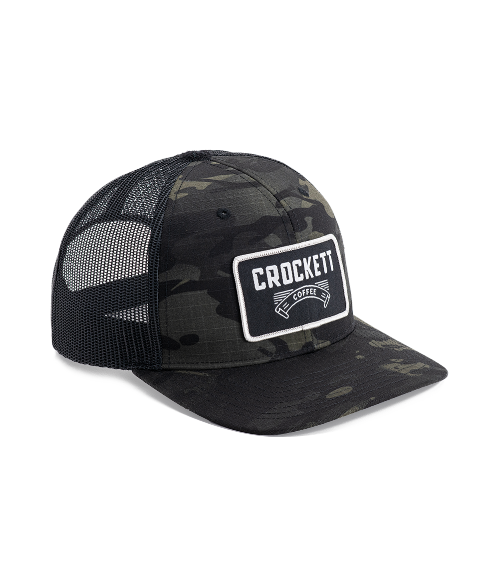 Black trucker hat with camo pattern, mesh back, and Crockett Coffee patch on the front.