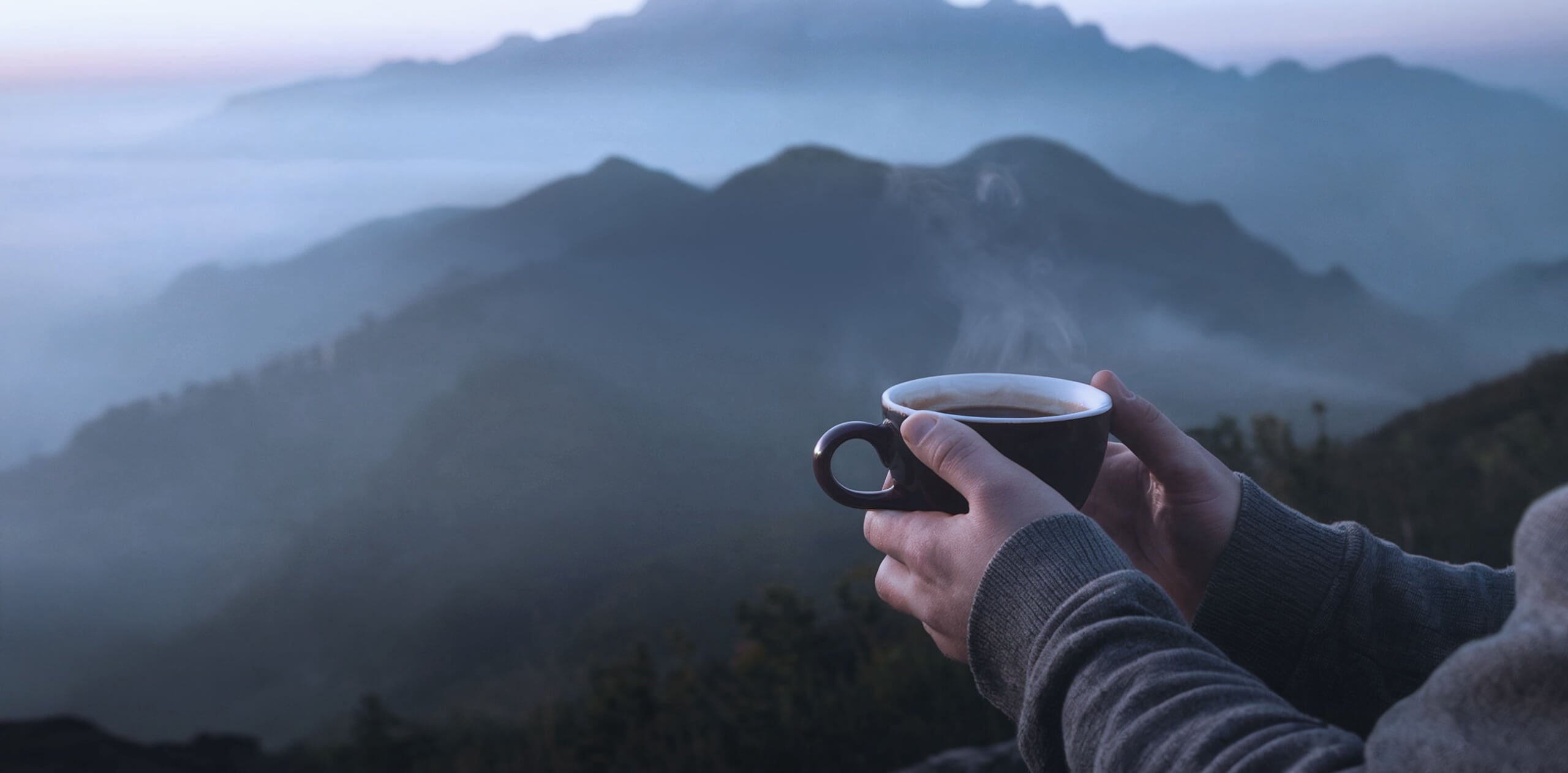 A person holding a steaming cup of coffee while overlooking misty mountains during sunrise.