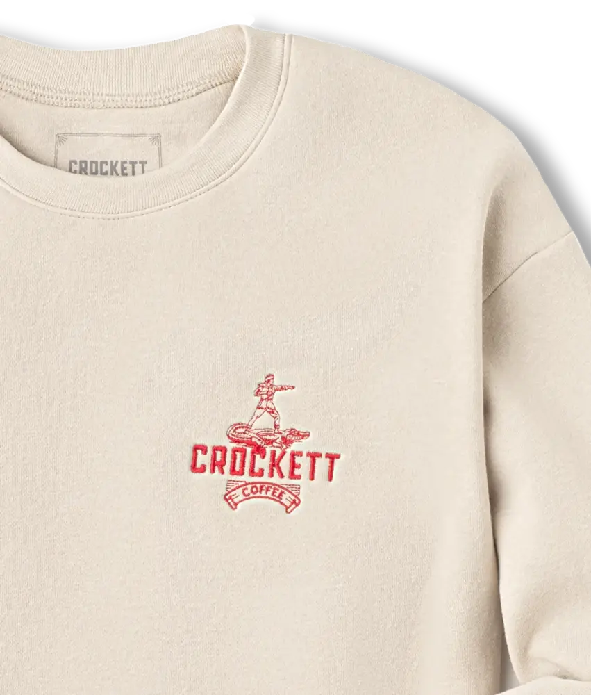 Off-white sweater with red embroidered Crockett Coffee logo and figure design on the left chest.