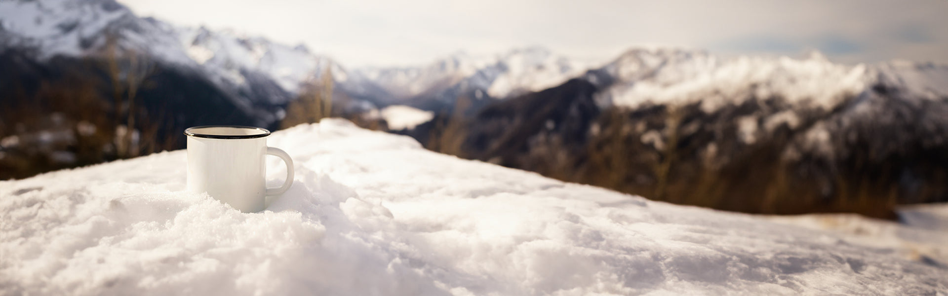 Coffee mug resting on snow with a scenic backdrop of snow-covered mountains and a clear sky.